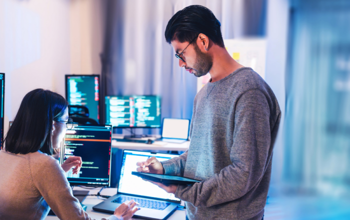 Two software developers collaborate in a bright office, one typing at a laptop while the other uses a tablet for code review.