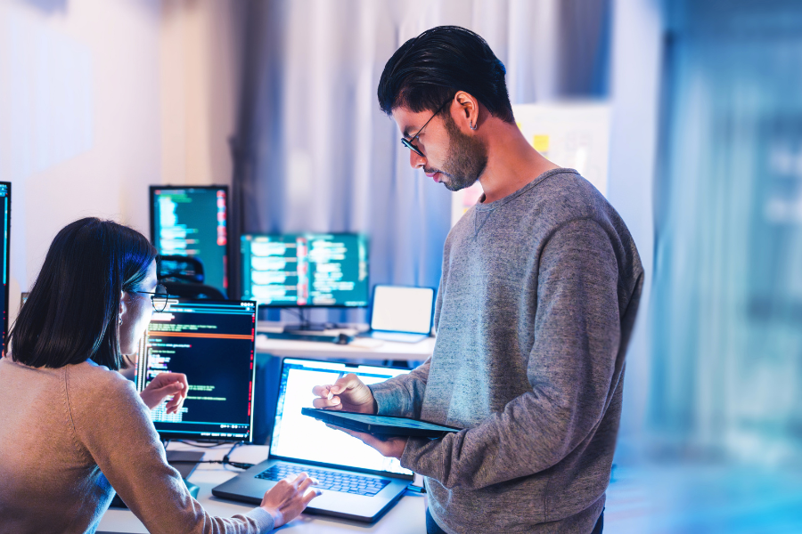 Two software developers collaborate in a bright office, one typing at a laptop while the other uses a tablet for code review.